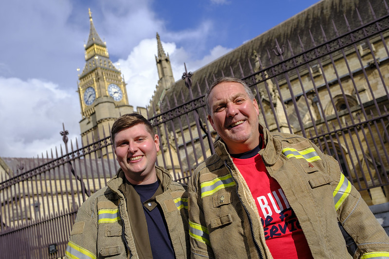 John Hodgkiss (right) and Matthew Wilkins (left) smile at camera wearing rebuild our fire service t shirts in front of houses of parliament