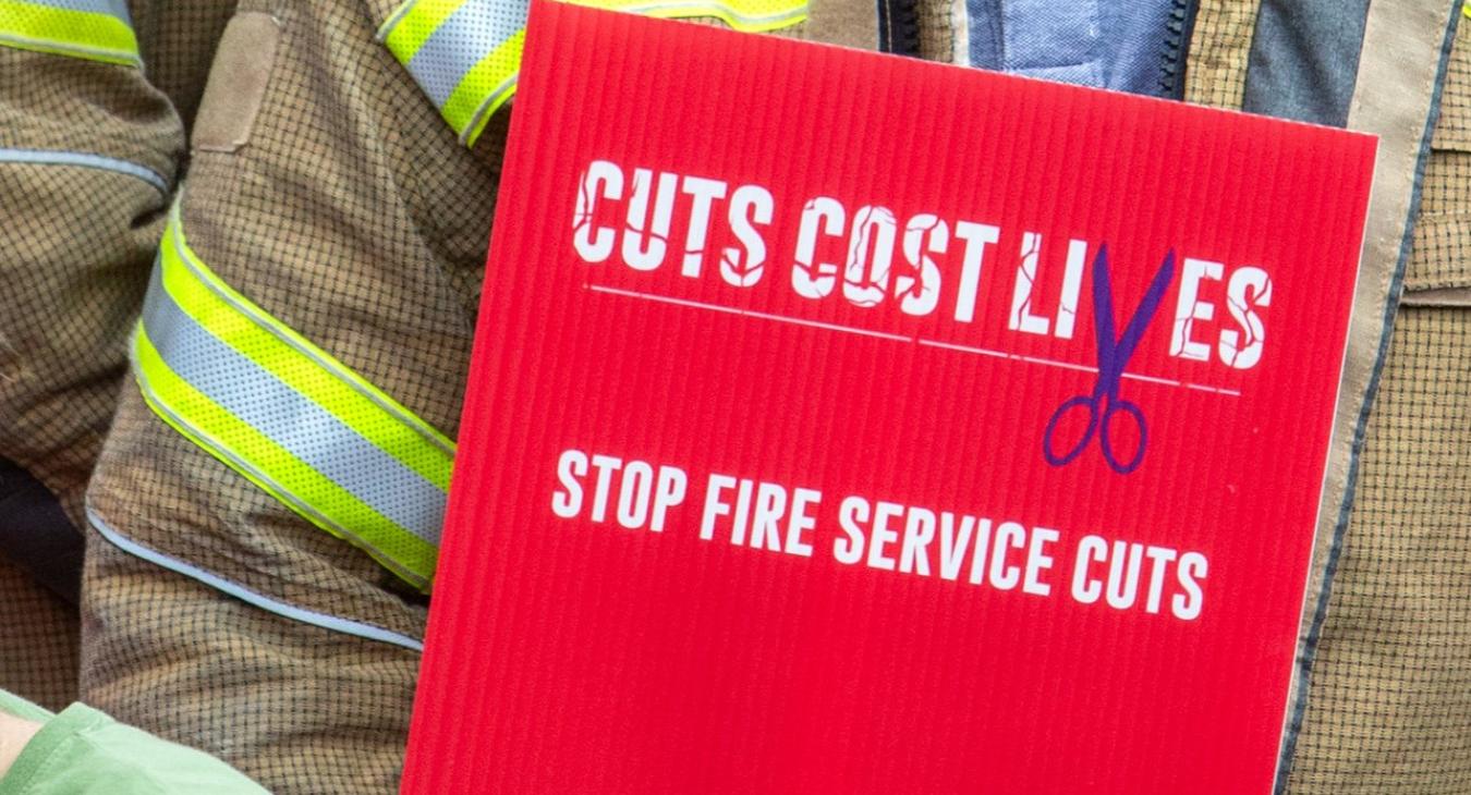 A firefighter holds a sign reading 'Cuts cost lives, stop fire service cuts'